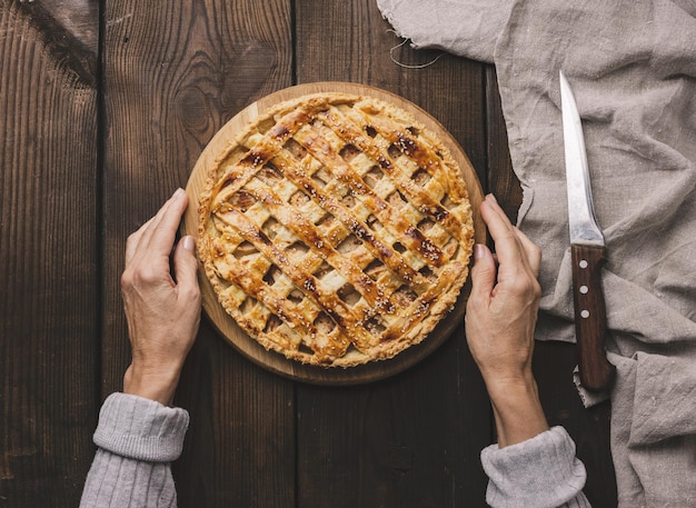 Zwei weibliche Hände halten einen runden gebackenen Kuchen mit Apfelfüllung auf einem braunen Tisch aus Holzbrett