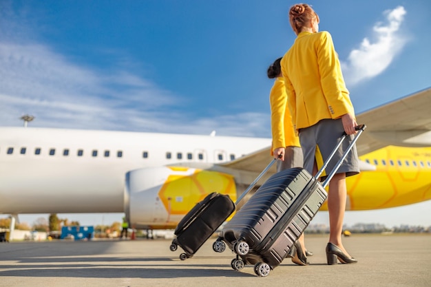 Zwei weibliche Stewardessen tragen Trolley-Gepäckkoffer am Flughafen mit Passagierflugzeug im Hintergrund