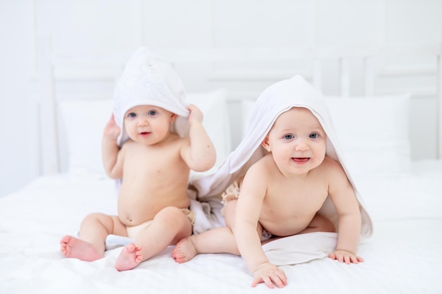Zwei Zwillingsbabys in einem weißen Handtuch nach dem Baden in einer Badewanne auf einem hellen Bett zu Hause lächeln das Konzept von Hygiene und Waschen