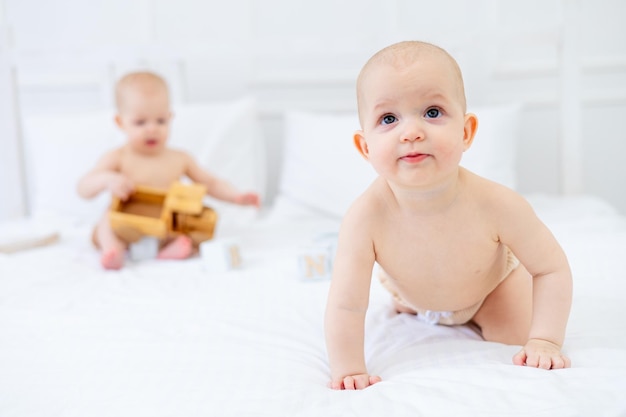 Zwei Zwillingsbabys von sechs Monaten in Windeln auf einem weißen Baumwollbett in einem Bodysuit auf einem Bett zu Hause, das mit Holzspielzeug spielt und lächelt