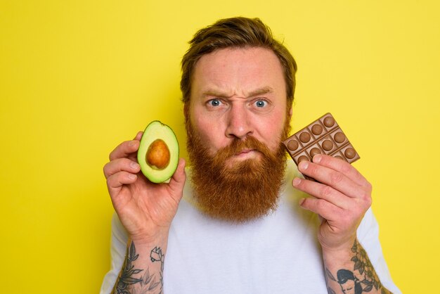 Zweiflermann mit Bart und Tattoos hält Avocado und Schokolade