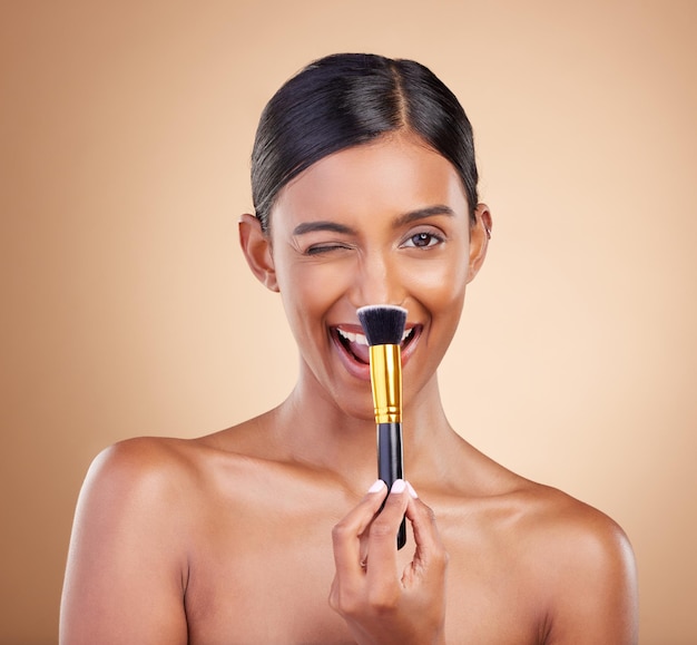 Zwinkernder Pinsel oder Make-up-Mädchen mit Beauty-Kosmetikprodukten oder Selbstpflege auf Studiohintergrund. Flirten Sie ein glückliches Modelgesicht oder eine junge Inderin mit Luxuskosmetik, strahlender Hautpflege oder einem Gesichtslächeln