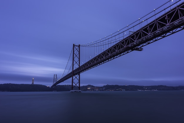 저녁에는 언덕과 빛으로 둘러싸인 Tagus 강 위의 25 de Abril Bridge