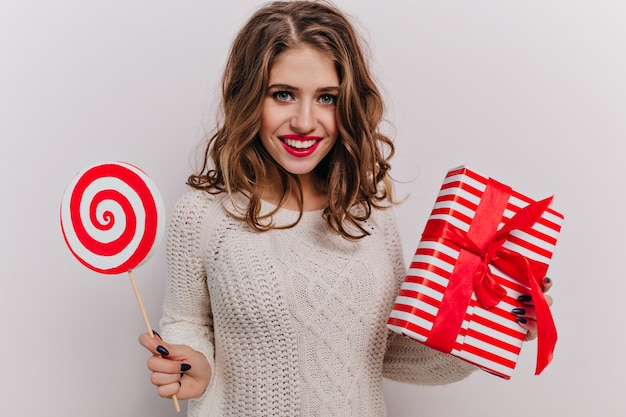 25 years old lady dressed in warm winter outfit with red lips and gorgeous eyelashes holding Christmas gift in red box with ribbon. Portrait of happy brunette with long curls