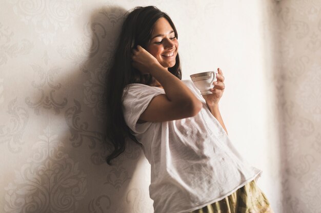 コーヒーカップを手にして壁紙に傾いている笑顔の女の子 無料の写真