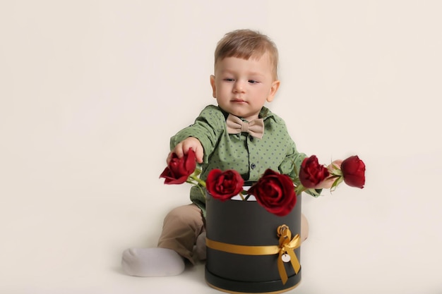 Красивый маленький мальчик в зеленой рубашке сидит на белом фоне рядом с коробкой с красными розами