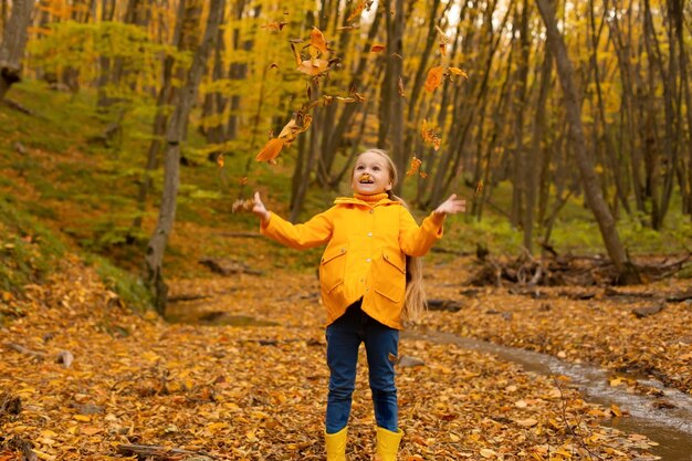 Красивая маленькая девочка в желтой куртке и сапогах гуляет по городскому осеннему лесу