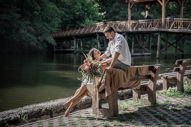 꽃을 든 아름다운 젊은 여성과 남편이 벤치에 앉아 의사소통, 자연에서의 데이트, 결혼의 로맨스를 즐기고 있습니다.