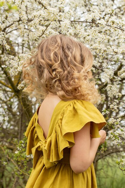 Блондинка в льняном платье стоит в весеннем цветущем саду и смотрит на цветы Premium Фотографии