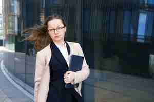Бесплатное фото Деловая женщина стоит с ноутбуком в костюме и очках возле офисного здания днем.