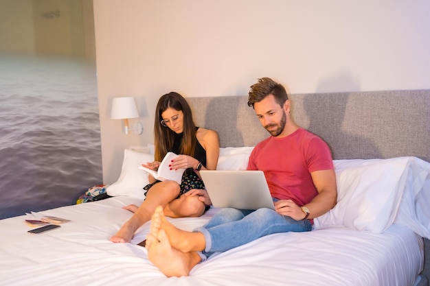 호텔 침대에 누워 컴퓨터와 책을 보고 있는 백인 부부