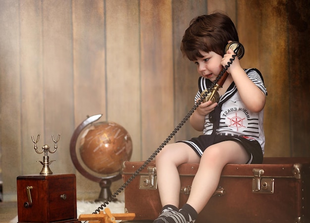 На полу сидит ребенок в ретро-интерьере и старый телефон. маленький ребенок путешественник в старинных украшениях. ребенок-путешественник звонит по телефону. Premium Фотографии
