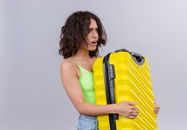 緑のクロップトップで短い髪の混乱した若い女性は、白い背景に黄色のスーツケースを考えて保持しています