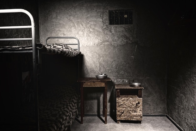 Темная пустая тюремная камера с двухъярусной кроватью и тумбочкой с алюминиевой посудой.