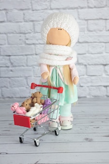 인형 소녀가 장난감과 플러시 천으로 된 다채로운 곰이 든 쇼핑 카트를 들고 서 있습니다.