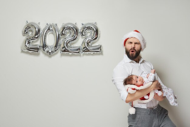 빨간 벨벳 산타 모자를 쓴 아버지가 2022년 모양의 은색 풍선 근처에서 어린 딸을 안고 있습니다. 어린 아이와 함께 수염 난 아빠가 신년 파티에서 놀란다.