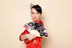 無料写真 伝統的な赤い和服で美しい日本の芸者のヘアスティッククリーム色の背景の日本でエレガントな扇子を保持しているポーズで正面