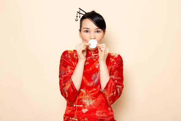 Бесплатное фото Вид спереди красивая японская гейша в традиционном красном японском платье с заколками для волос, держащей маленькое белое выражение хлопка на кремовом фоне церемонии японии