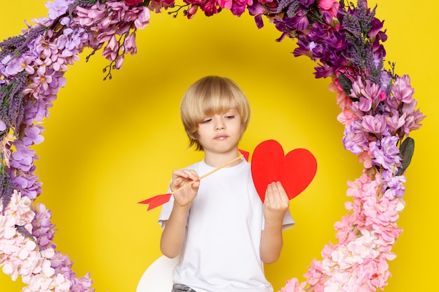 Бесплатное фото Вид спереди blodne kid в белой футболке, держащей сердечко, сидящей на цветочной подставке на желтом пространстве