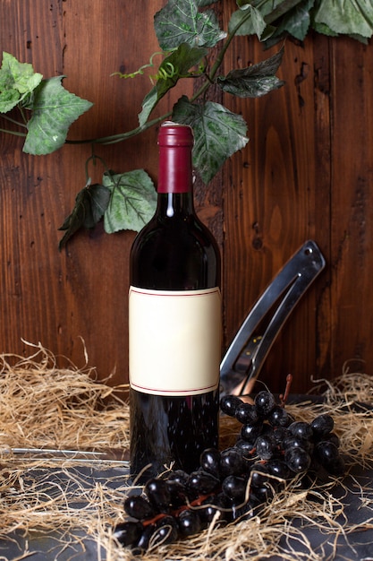 Фронтальный вид бутылки алкоголя в черной бутылке с бордовым колпачком вместе с черным виноградом и зелеными листьями на коричневом фоне пьет винзавод алкоголь
