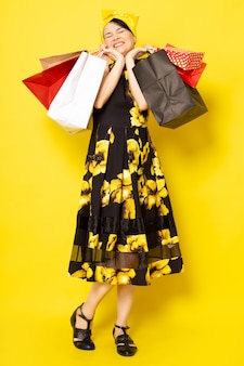 黄色​の​黒い​花​の​正面​の​若い​魅力的​な​女性​は​、​黄色​の​ショッピング​パッケージ​を​保持している​ポーズ​の​頭​の​上に​黄色​の​包帯​で​ドレス​を​デザインしました