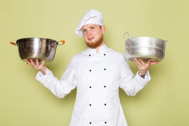 Бесплатное фото Вид спереди молодого мужчины-повара в белом кухонном костюме. белая шапка с серебряными и металлическими кастрюлями.