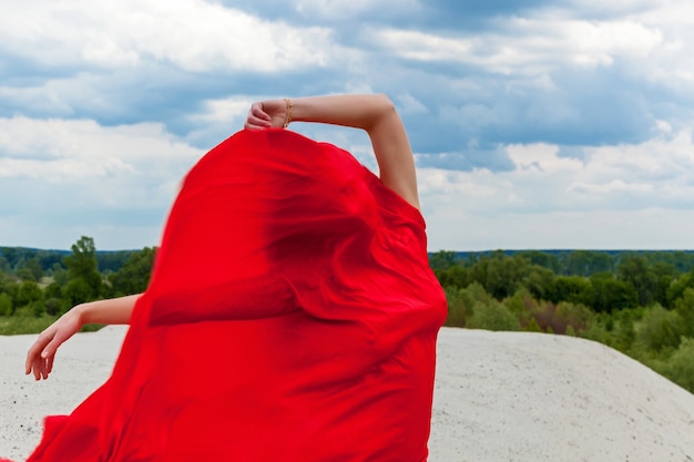 砂の上の赤い布の女の子は、曇り空を背景に写真家のポーズをとります。風に吹かれた赤い布が少女の姿を抱きしめます。
