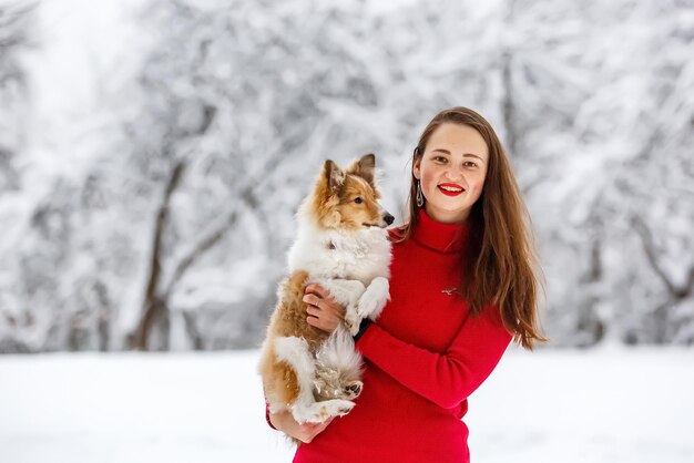 Девушка в красном платье с собакой шелти на руках. зимний фон.