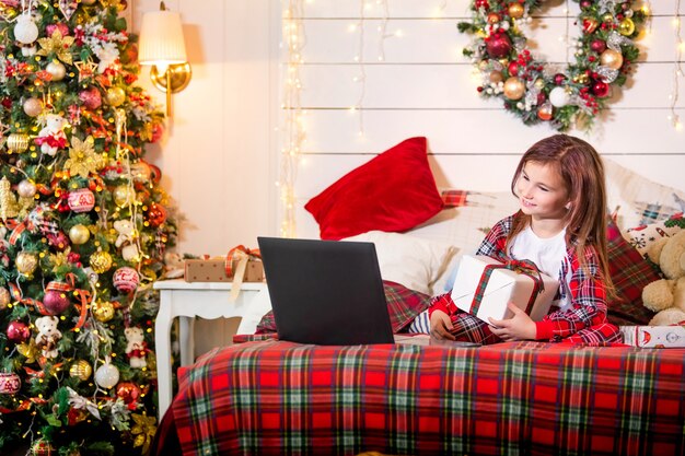 パジャマ姿の女の子は クリスマスツリーの赤い格子縞のベッドに横たわって ラップトップのモニターを見ています プレミアム写真