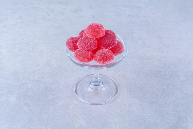 Стеклянная тарелка, полная красных сладких желейных конфет на сером столе.