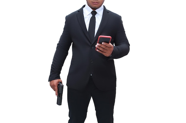 黒のスーツを着た銃の男がピストルを持っており、もう一方の手は白い背景の孤立したミッションでターゲットをチェックするために彼の携帯電話に向かっています。暗殺者、殺人、犯罪者、泥棒のコンセプト。