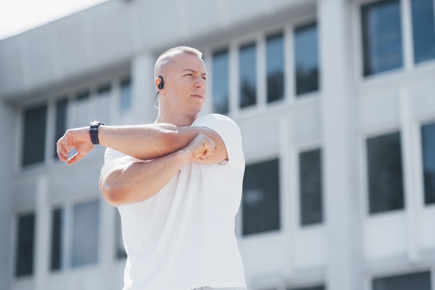 Красивый фитнес-мужчина в спортивной одежде, делает растяжку во время подготовки к серьезным упражнениям в современном городе