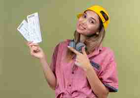 Бесплатное фото Счастливая молодая женщина в красной рубашке и желтой бейсболке с наушниками, указывая на билеты на самолет указательным пальцем на зеленой стене