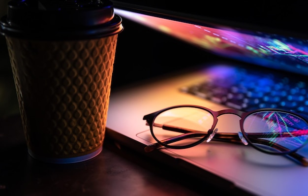 Бесплатное фото Ноутбук, наполовину закрытый в темноте, с красочной светящейся чашкой кофе и стаканами