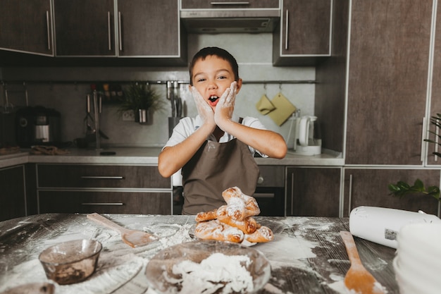 Маленький мальчик дома на кухне лепит на столе тесто и смеется. малыш радуется, что его испачкали в муке.