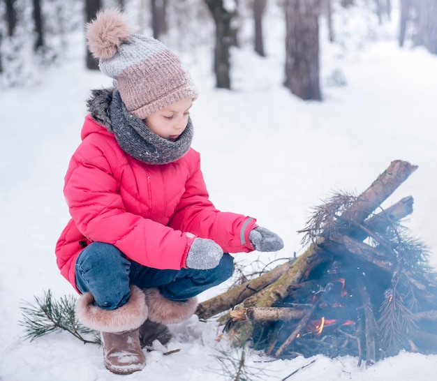 밝은 재킷을 입은 어린 소녀가 겨울 눈 덮인 숲에서 놀고 있습니다. 소녀는 불에 의해 워밍업됩니다.