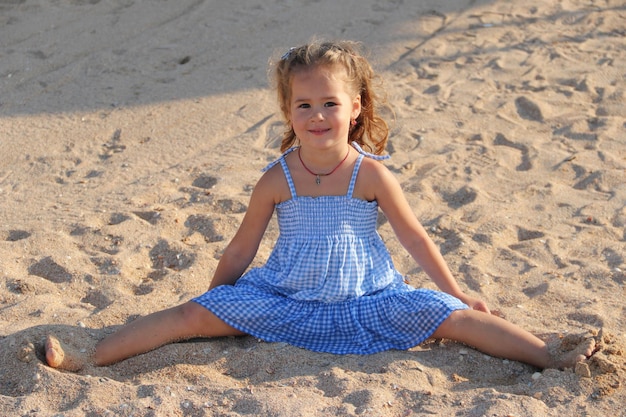 Маленькая девочка сидит на песке в шпагате в голубом платье концепция детской радости