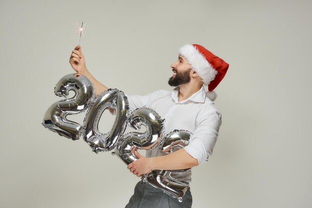 빨간 벨벳 산타 모자를 쓴 남자가 한 손에는 2022년 모양의 은색 풍선을, 다른 한 손에는 불꽃을 들고 있습니다. 수염을 기른 행복한 남자가 신년 파티에서 폭죽을 보고 웃고 있습니다.