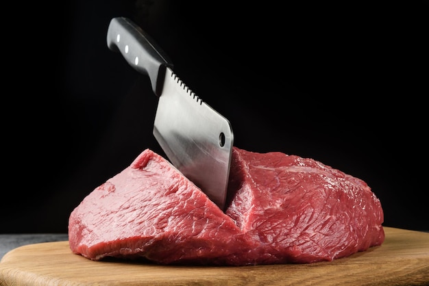Кусок сырого мяса крупным планом с топором шеф-повара на темном фоне.