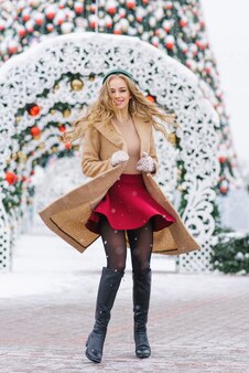 Игривая женщина в бежевом пальто и бордовой юбке кружится и танцует на городской улице, наслаждаясь зимними праздниками на рождественской ярмарке. концепция городского образа жизни