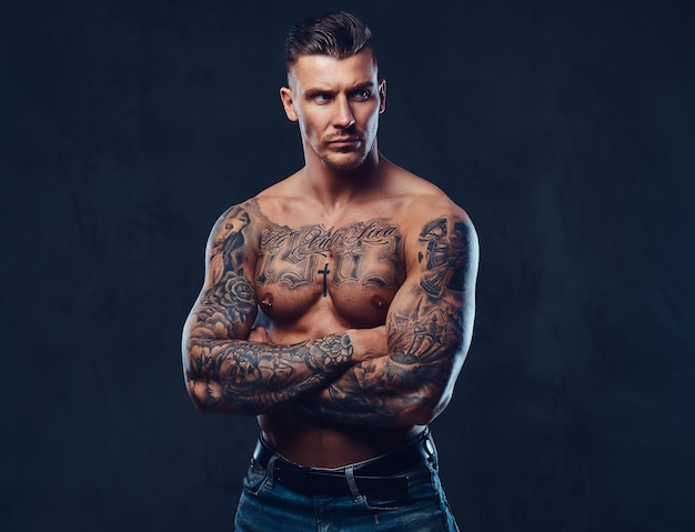 Бесплатное фото Татуированный мускулистый мужчина без рубашки со стильными волосами позирует перед камерой на темном фоне.