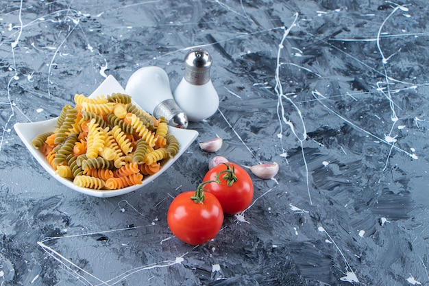 Белая миска, полная разноцветных макарон с овощами и специями на мраморной поверхности.
