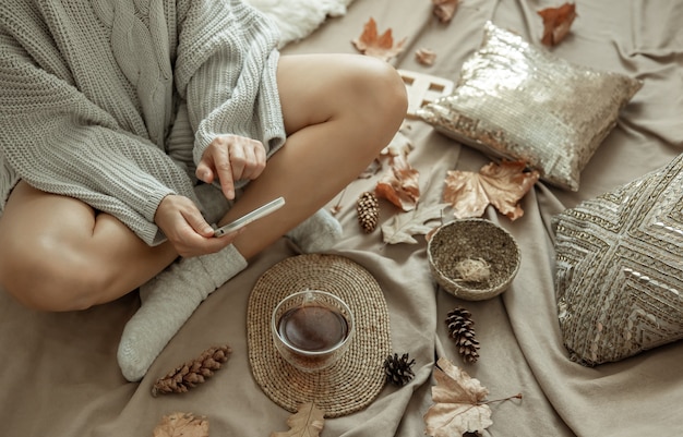 Бесплатное фото Женщина в вязаном свитере фотографирует чашку чая в постели среди шишек и листьев, осеннюю композицию, довольство.
