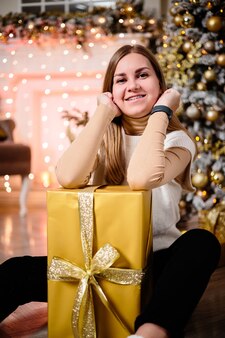 흰 스웨터를 입은 여성, 크리스마스 인테리어의 따뜻하고 아늑한 저녁, 조명, 선물, 장난감, 화환으로 장식된 크리스마스 트리. 축제의 거실. 새해