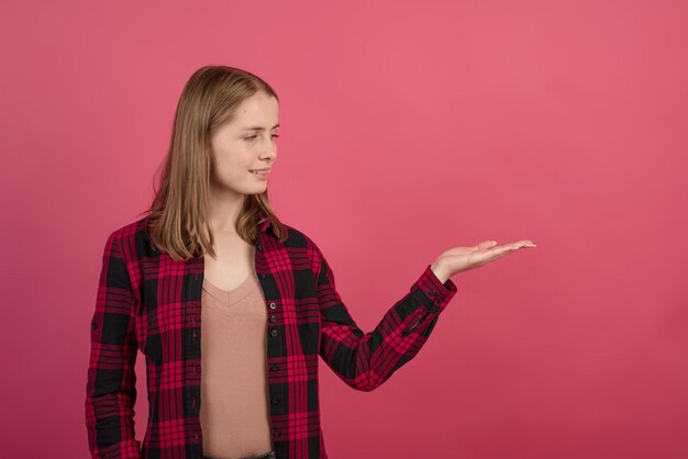 ピンク​の​スタジオ​の​背景​に​広告​の​ために​手のひら​に​空き​スペース​を​保持している​シャツ​を​着た​若くて​魅力的​な​金髪​の​白人​の​女の子​。