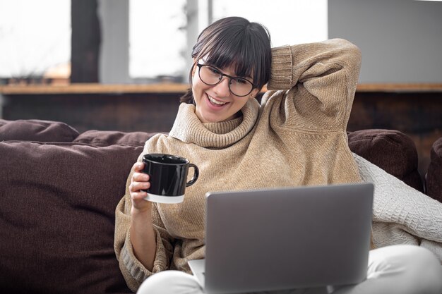Молодая красивая девушка сидит дома на диване с чашкой напитка и весело смотрит на экран компьютера. понятие видеосвязи и общения на расстоянии.