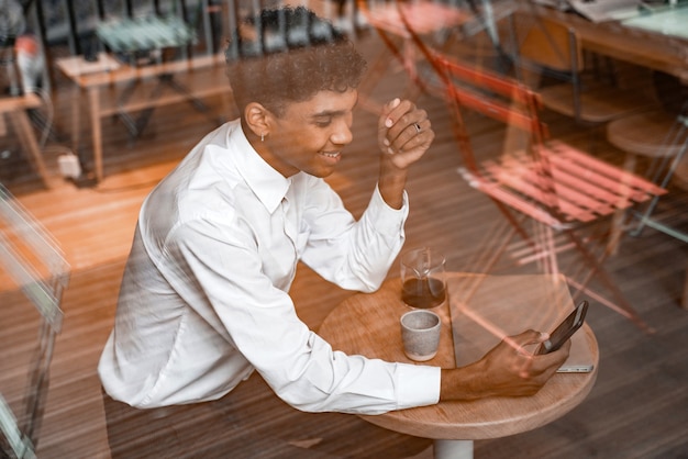 한 젊은 흑인 남성이 노트북을 들고 탁자에 앉아 차를 마시고 스마트폰을 사용하고 있습니다. 카페의 그 남자. 사무실 밖에서 일하거나 원격 작업 또는 연구 개념.