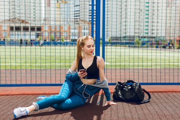 Бесплатное фото Молодая девушка в синем спортивном костюме с черным верхом сидит у забора на стадионе. она слушает музыку в наушниках и смотрит вдаль.