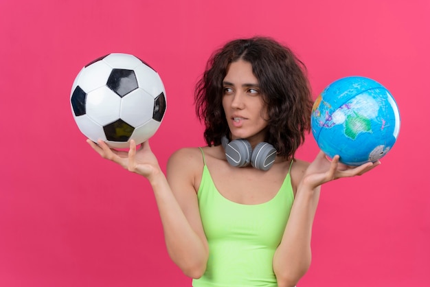 Бесплатное фото Молодая красивая женщина с короткими волосами в зеленом топе в наушниках держит глобус и смотрит на футбольный мяч