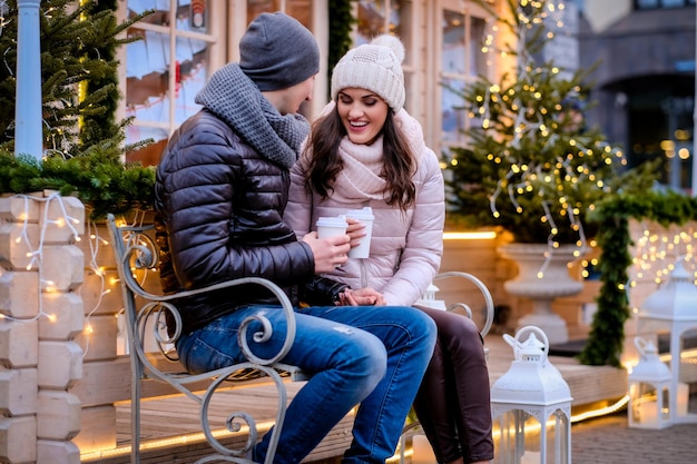 無料写真 美しいライトで飾られたイブニングストリートのベンチに座って、クリスマスの時期に屋外でコーヒーを飲みながら話したり、暖めたりして、暖かい服を着ている若いロマンチックなカップル